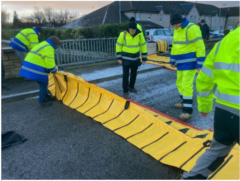 A group of people in yellow jackets trialling out the rolling out of flexible flood barriers.